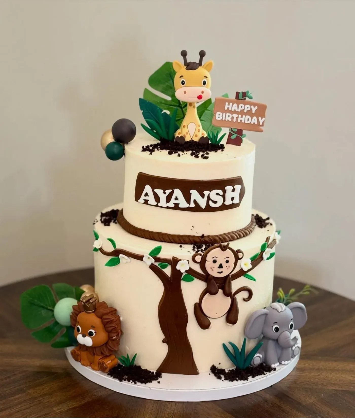 Two-tier cream birthday cake decorated with a hand-modeled giraffe, monkey swinging from a painted tree, lion and elephant, finished with chocolate-soil detail and green foliage.