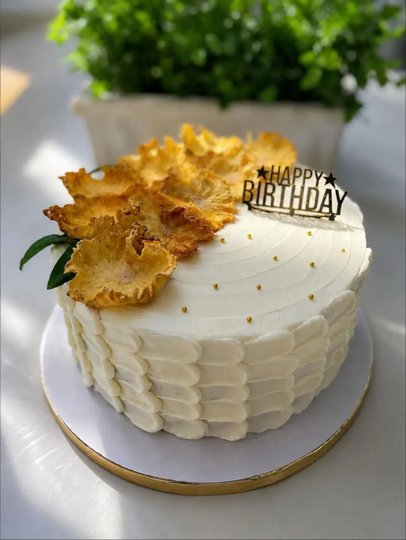 White buttercream cake with a scalloped palette-knife finish, crowned with dried pineapple flowers, gold dragees and a black 'Happy Birthday' topper.