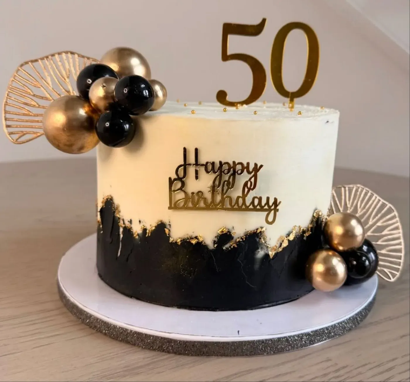 Cake split between ivory buttercream and a torn black band edged in gold leaf, dressed with black and gold balloon spheres, gold fans and a gold '50' topper.