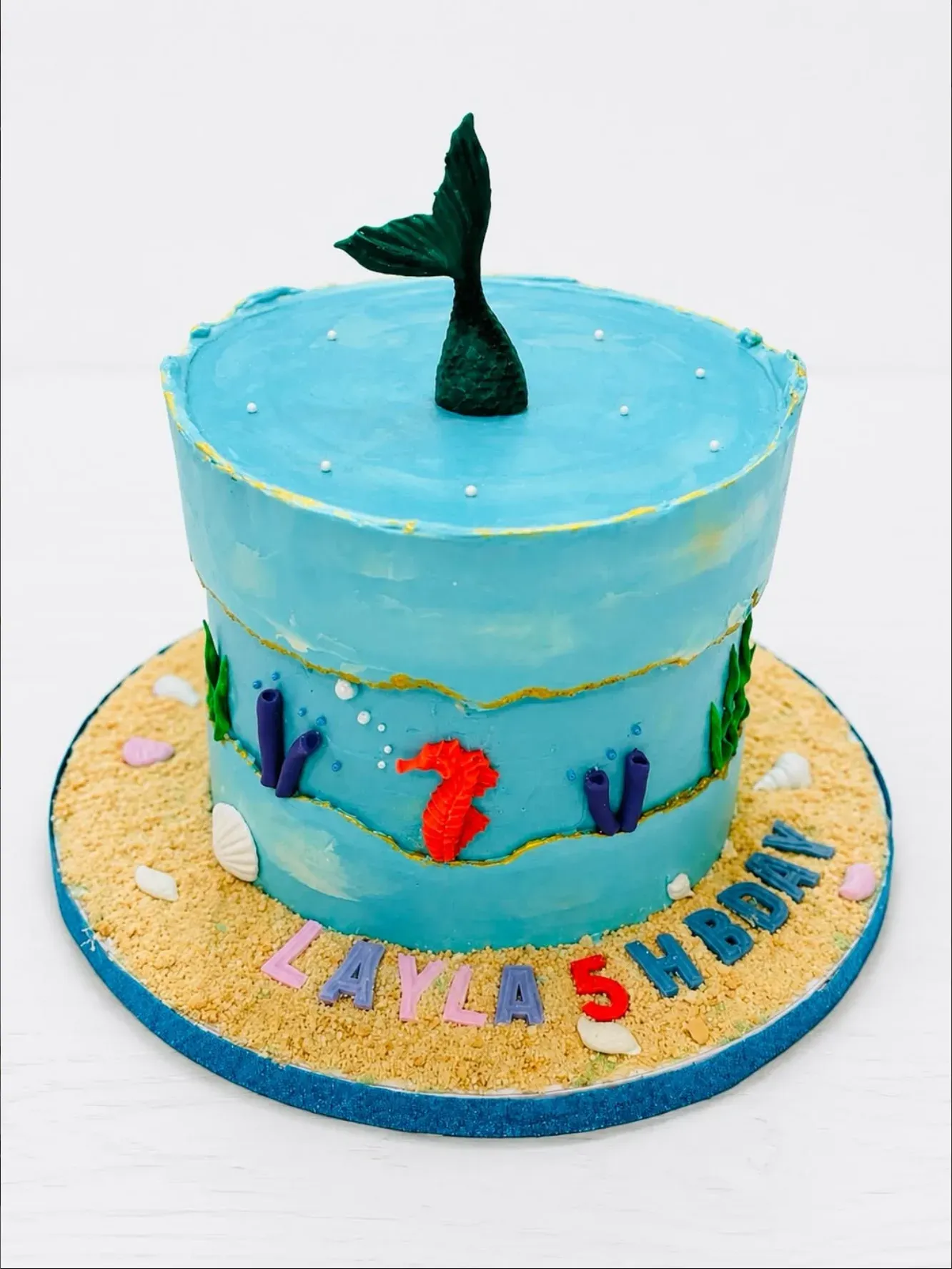 Turquoise ocean-effect cake with a mermaid tail rising from the top, piped seaweed, a seahorse and shells, set on a biscuit-crumb sand board.