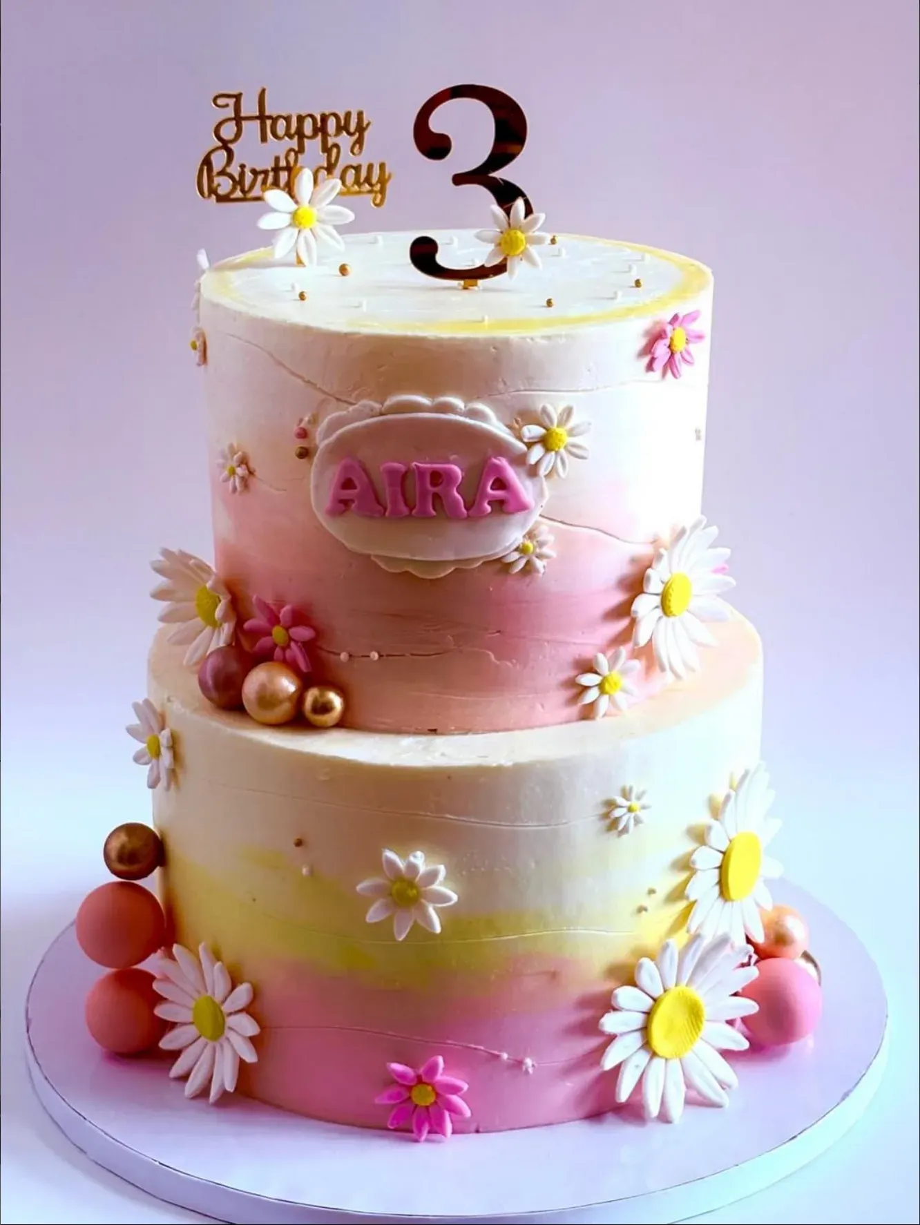 Two-tier cake washed in pink, peach and yellow ombré buttercream, covered in sugar daisies and blossom with gold and coral spheres at the base.