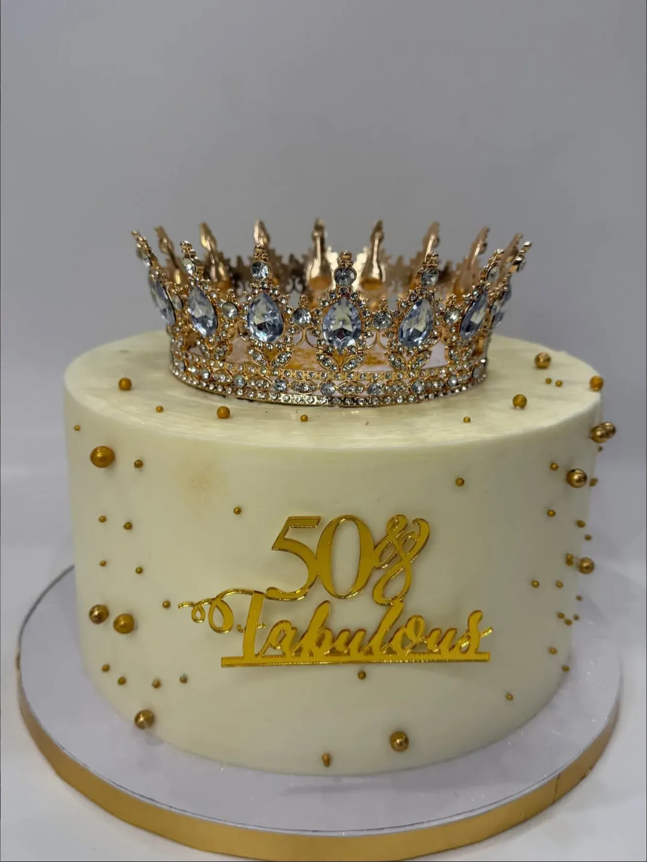Ivory buttercream cake topped with a jeweled gold crown, scattered with gold pearls and a gold '50 & Fabulous' plaque.