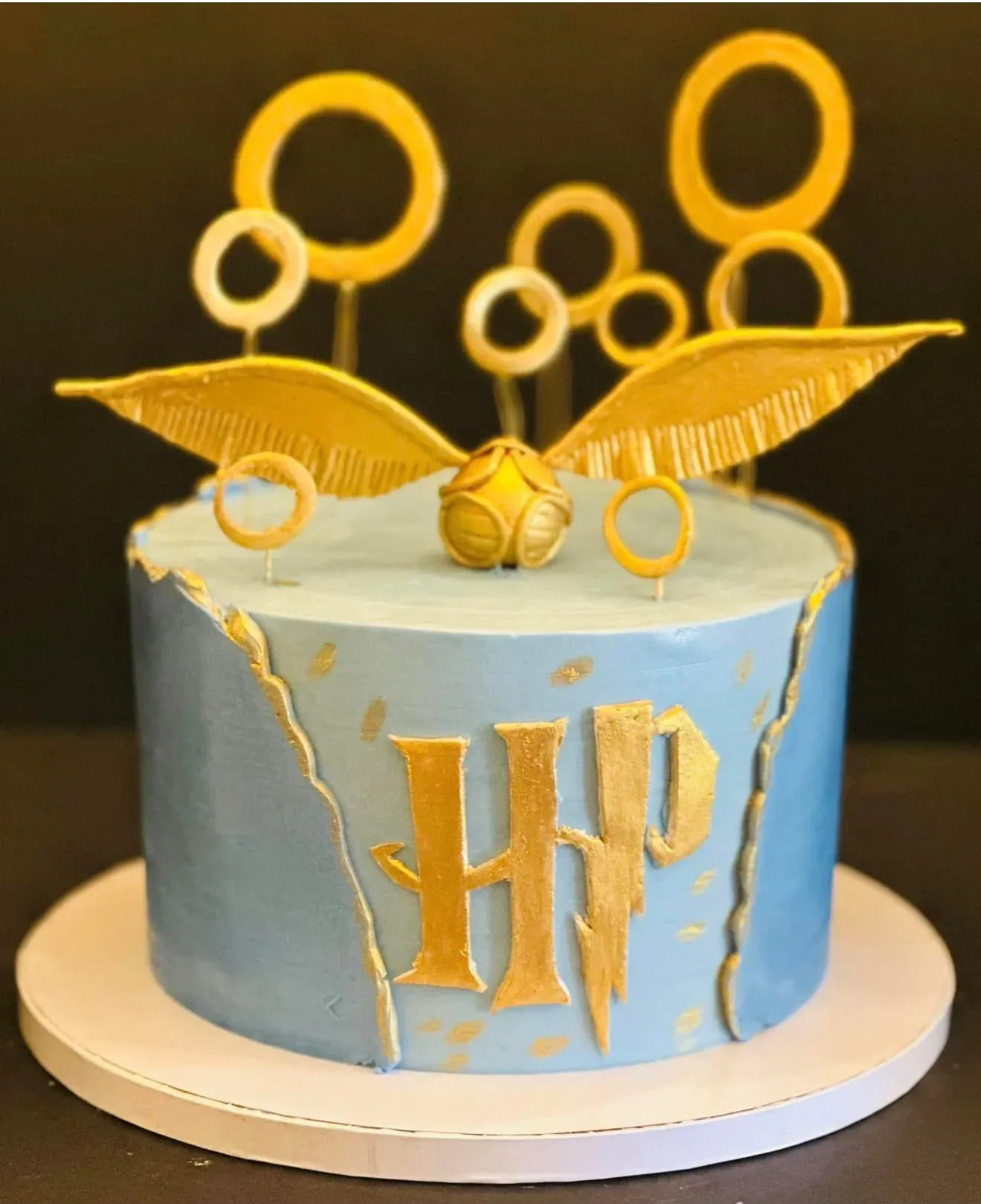 Dusty blue cake with a torn gold-leaf seam, a gold monogram on the side, topped with a winged golden snitch and a halo of gold wire rings.