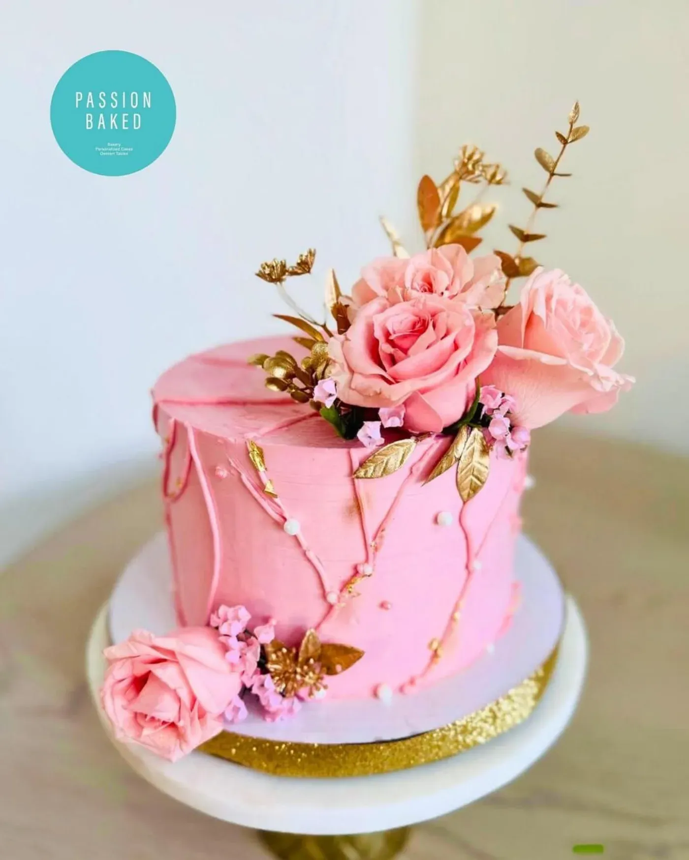 Single-tier pink buttercream cake with a textured palette-knife finish, gold leaf detailing and a crown of fresh pink roses and blossom.