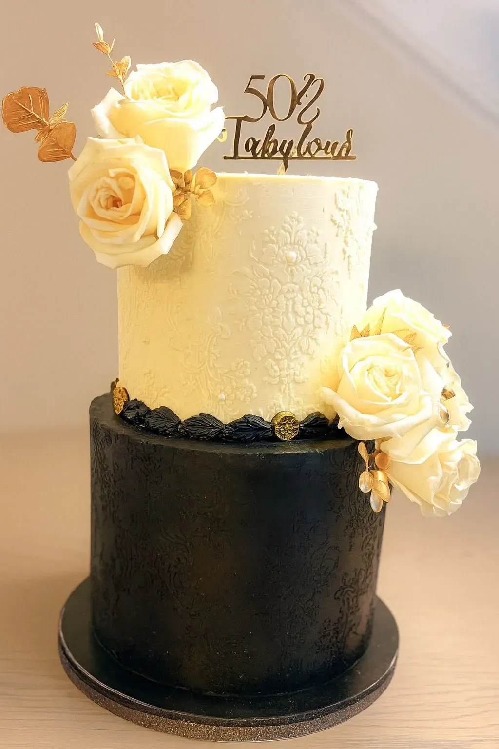 Two-tier cake with a textured black lower tier and an ivory upper tier embossed with lace, dressed with cream garden roses, gold leaves and a gold 'fabulous' topper.