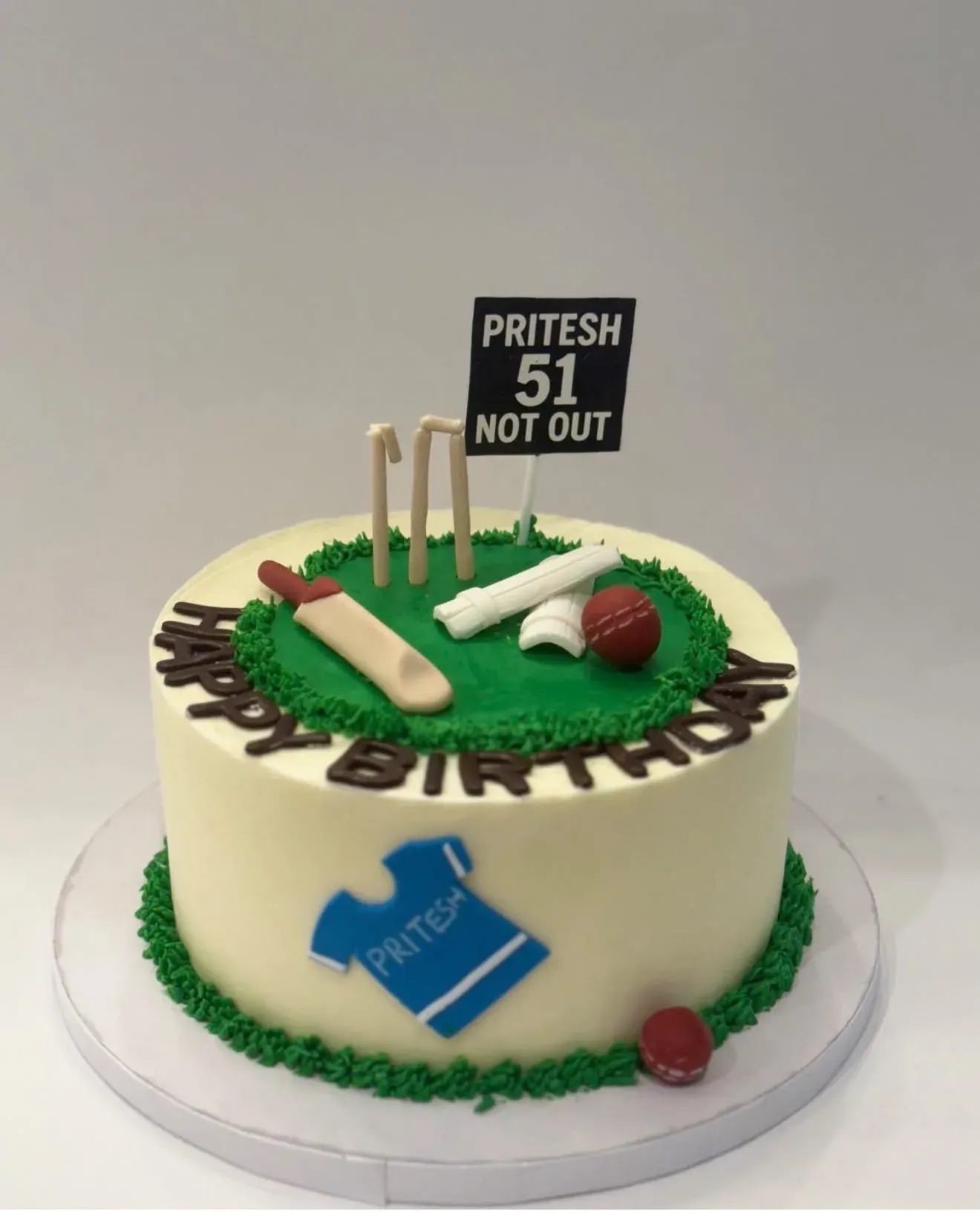 Cricket-themed birthday cake with a piped green outfield, sugar bat, stumps and ball on top, a fondant team jersey on the side and a black scoreboard topper reading 'not out'.