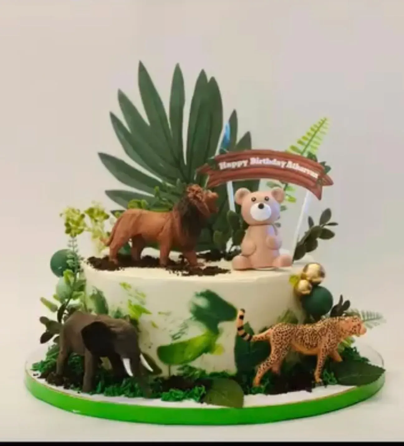Buttercream cake with a green watercolor wash, crowned with sugar palm leaves and ferns and a group of modeled safari animals — lion, elephant, leopard and a seated bear — with a banner topper.