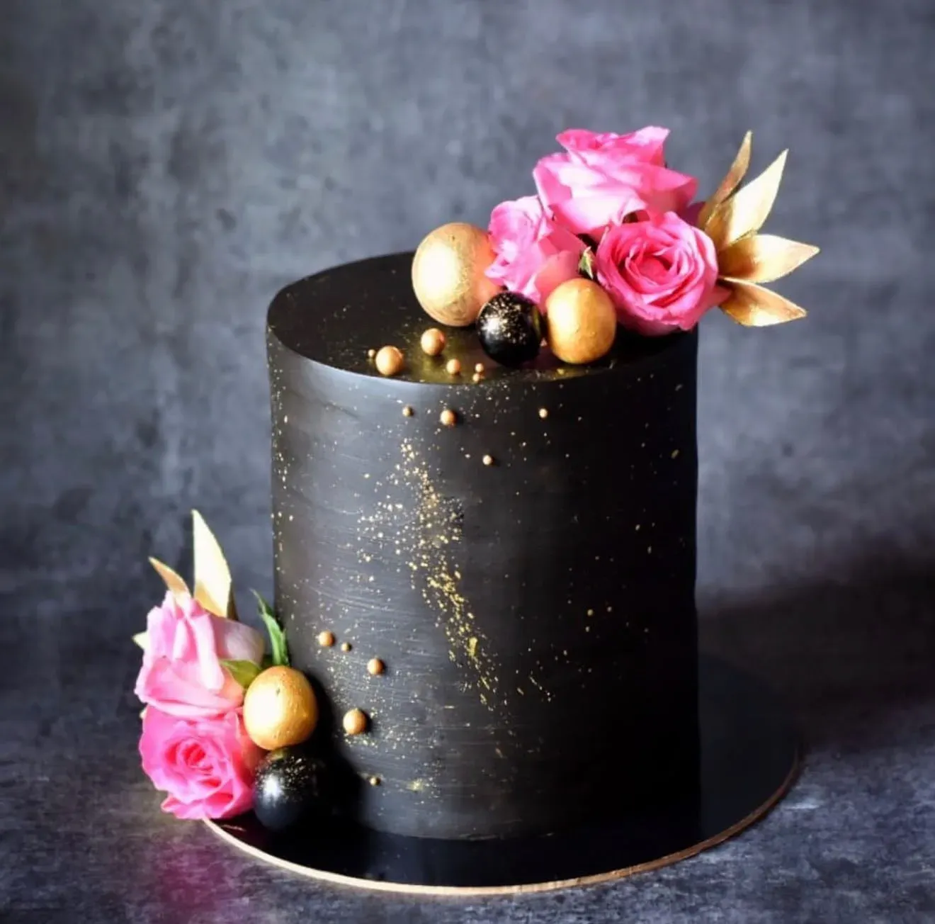 Tall matte black cake flecked with gold dust, trimmed with hot pink roses, gold and black spheres and gold sugar leaves.