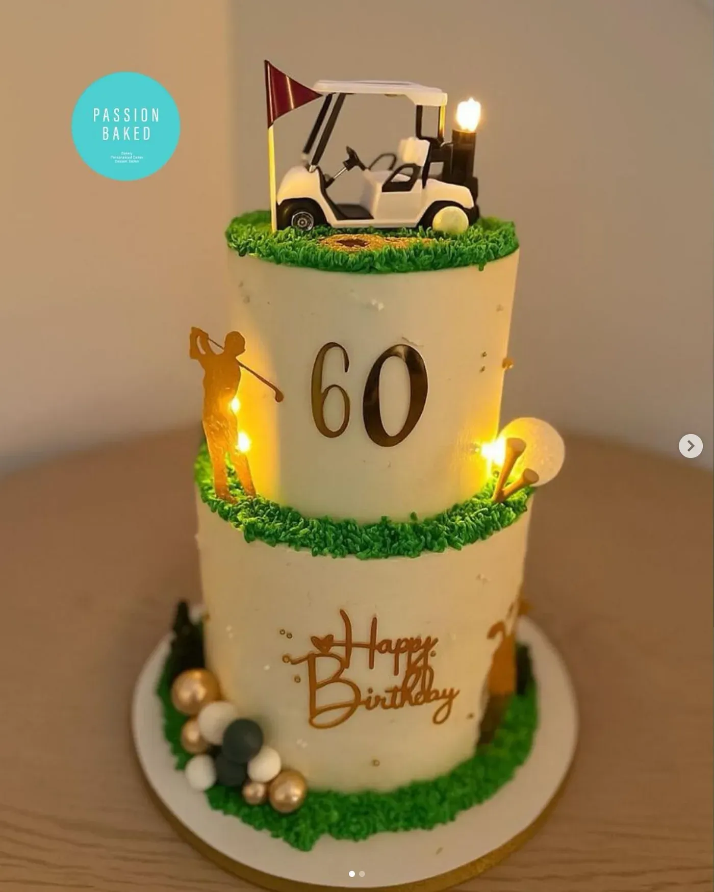 Two-tier ivory cake with piped grass edging, a gold golfer silhouette and '60' on the upper tier, topped with a golf buggy and flag, lit with warm fairy lights.