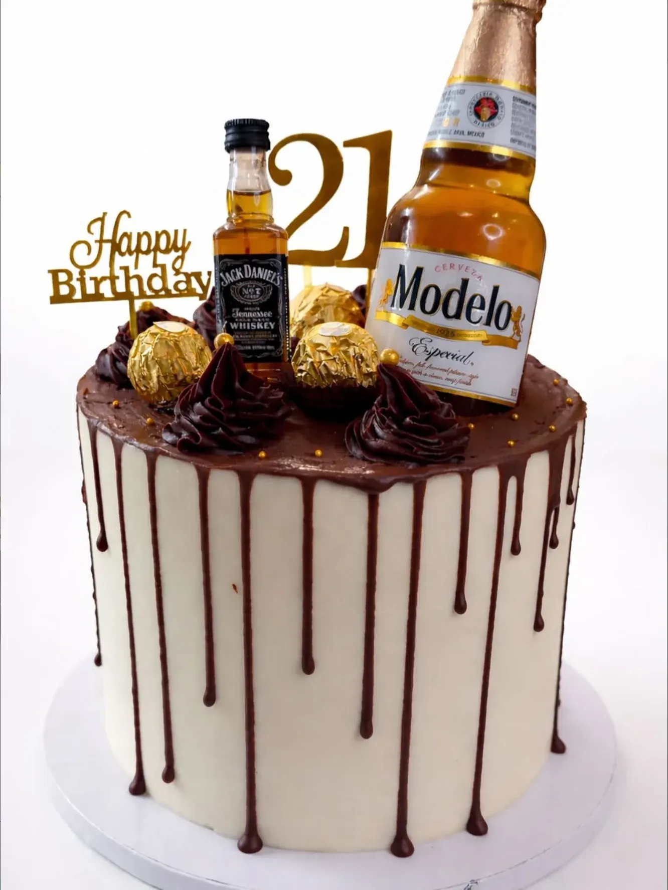 Ivory buttercream cake with a dark chocolate drip, piped ganache swirls and gold-wrapped chocolates, topped with miniature bottles and a gold '21'.