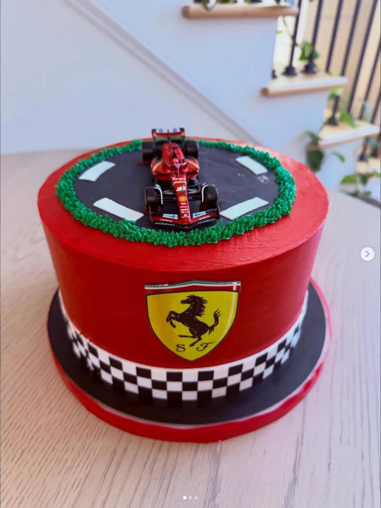 Red cake with a piped racetrack and grass on top carrying a model Formula One car, wrapped with a black and white checkered band and a racing crest.
