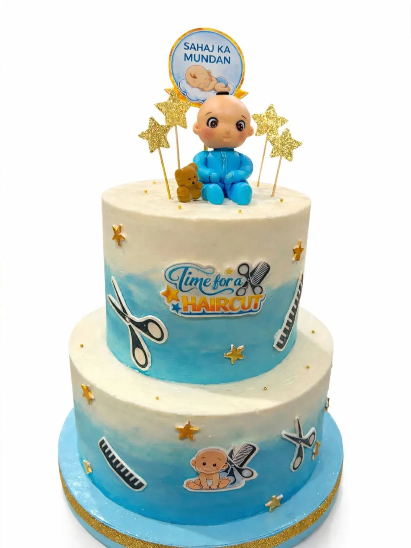 Two-tier cake in blue and white ombré buttercream with gold stars, scissors and comb motifs, topped with a modeled baby figure and glitter stars.