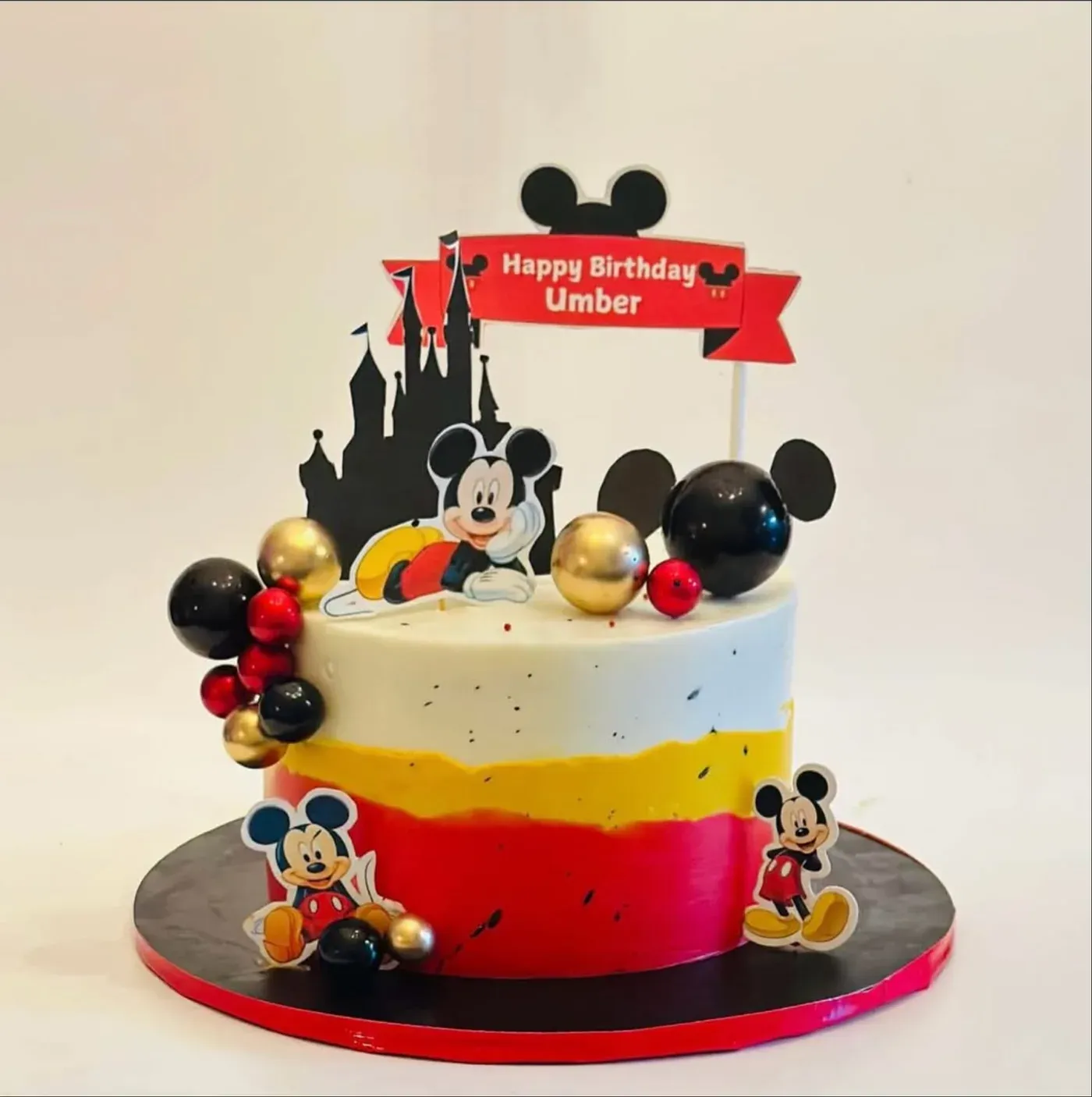 Red, yellow and white layered buttercream cake with a castle and Mickey Mouse topper, finished with black, red and gold balloon spheres.