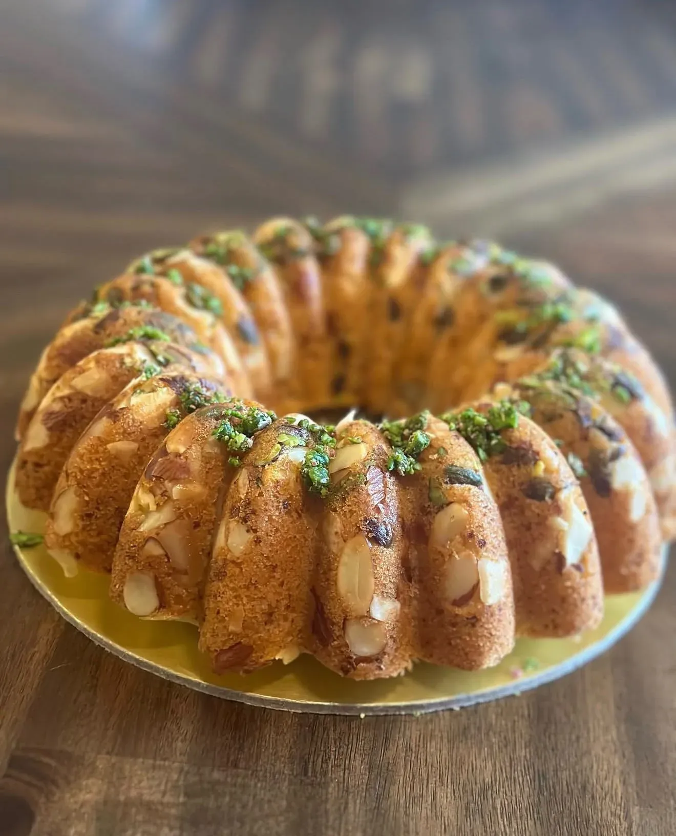 Golden bundt-style tea cake studded with flaked almonds and scattered with chopped pistachios, served on a gold board.